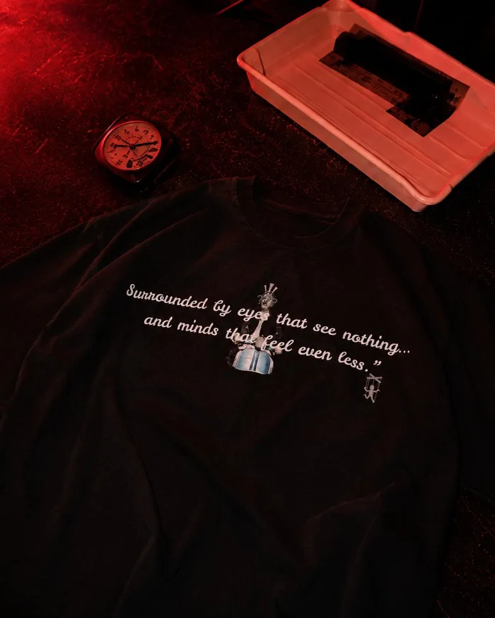 Black printed T-shirt laid flat beside a tray and clock in a red-lit room.